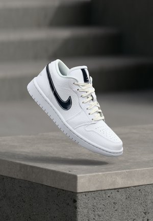 Zapatilla blanca de cuero con el logo negro de Nike, cordones atados, flotando sobre un borde de hormigón gris y escaleras con fondo desenfocado.