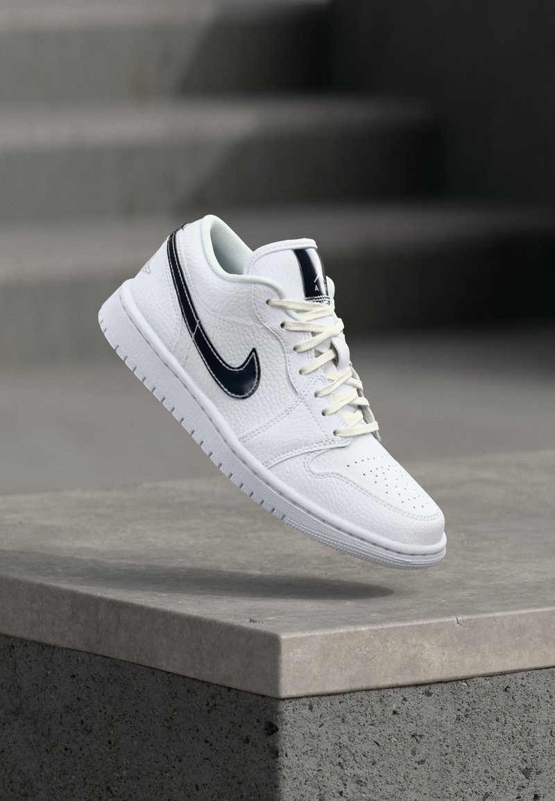 Λευκό δερμάτινο αθλητικό παπούτσι με μαύρο λογότυπο Nike, δεμένα κορδόνια, να αιωρείται πάνω από γκρι τσιμεντένιο περίβολο και σκάλες με θολό φόντο.