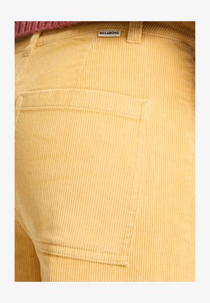 Geel corduroybroek met verticale ribbels, voorzien van een achterzak en een klein merketiket. De stof heeft een zachte textuur.