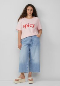 Lichtroze t-shirt met "spicy." in rode tekst, gecombineerd met lichtgewassen, wijduitlopende jeans met rafels en beigerode plateau-sandalen.