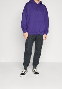 Felpa viola con cappuccio e tasca frontale a marsupio, abbinata a pantaloni cargo scuri con tasche laterali e orli risvoltati. Scarpe da ginnastica nere e bianche.