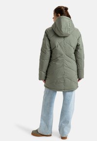 Doudoune olive avec capuche, présentant un motif en chevrons et des manches longues, associée à un jean large bleu clair et des bottes marron.