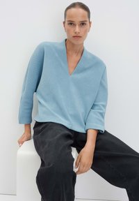 Lichtblauw, geribbelde blouse met een diepe V-hals en driekwartmouwen, gecombineerd met losse zwarte broek. Gladde textuur en casual ontwerp.