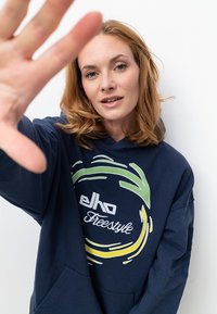 Marineblaue Kapuze aus weichem Stoff, mit einem grafischen Logo in Grün und Gelb. Das Design umfasst eine Vordertasche und eine lockere Passform.