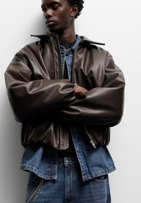 Hnedá kožená bomber bunda s voľným strihom, rebrovanými manžetami a zipsom vpredu, prenesená cez džínovú bundu, kombinovaná s modrými džínami.