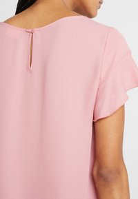 Blouse rose avec un col rond et de courtes manches volantées. Une ouverture en goutte d'eau au dos est maintenue par un bouton. Texture du tissu lisse.