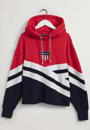 Haut à capuche rouge et marine avec des rayures blanches, présentant un logo avec un blason et le mot "BANT". Accroché à un cintre blanc sur un fond clair.