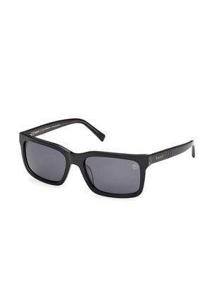 Gafas de sol negras en forma rectangular con lentes oscuros, montura de plástico grueso y un logotipo plateado en la patilla. Textura suave y diseño simple.