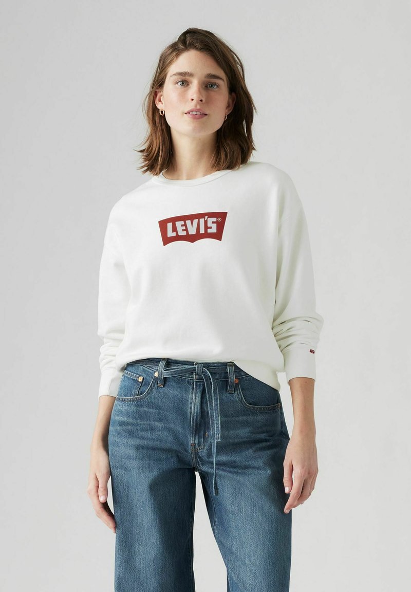 Vit sweatshirt med Levi's röda logotyp på bröstet, tillsammans med blå denimjeans med dragsko i midjan och avslappnad passform.