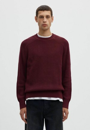 Un homme porte un pull bordeaux côtelé à col rond sur une chemise blanche et un jean foncé, sur un fond clair uni.