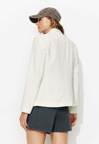 Blazer blanc au design structuré, doté de manches longues et d'une texture lisse, associé à un short gris foncé et une casquette imprimé léopard.