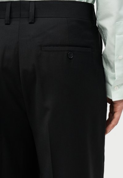 Κοντινό πλάνο ενός ατόμου που φοράει μαύρο παντελόνι με κουμπωμένη πίσω τσέπη και άσπρο πουκάμισο με μακριά μανίκια.