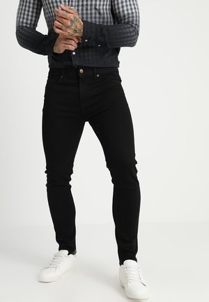 Mand iført sorte slim-fit jeans, en ternet sort og grå langærmet skjorte, hvide sneakers, med tatoverede hænder, der er foldet sammen.