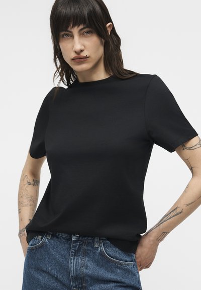 Γυναίκα με καστανά μαλλιά μέχρι τον ώμο, ντυμένη με απλό μαύρο κοντομάνικο μπλουζάκι και μπλε τζιν, με εμφανή τατουάζ και στα δύο χέρια.