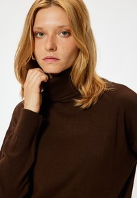Suéter de cuello alto marrón con textura acanalada, que presenta mangas largas y un cuello ajustado. La tela parece suave y cómoda.
