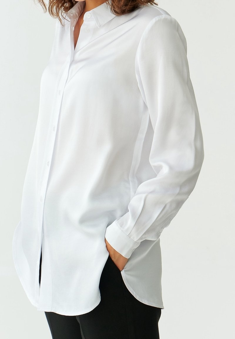 Femme portant une chemise blanche à manches longues avec col, rentrée derrière une main dans un pantalon noir, sur un fond clair uni.