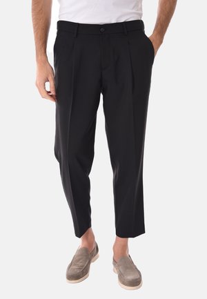 Uomo che indossa pantaloni neri pieghettati corti, camicia bianca e scarpe slip-on in camoscio grigio, in piedi con una mano in tasca.