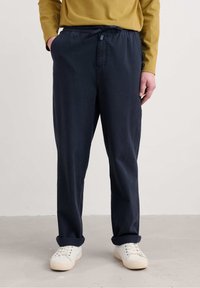 Pantaloni din bumbac bleu marin cu un brâu elastic și șnur ajustabil. Au buzunare laterale și manșete răsucite. Purtați cu adidași albi.