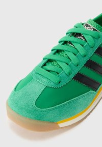 Sapatilha desportiva verde com um upper em camurça e malha, riscas pretas, atacadores texturizados e uma sola em borracha de goma com detalhes amarelos.