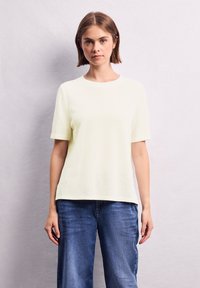 Lichtgeel topje met korte mouwen, een ronde hals, textuurstof en een losse pasvorm, gecombineerd met blauwe jeans met brede pijpen.