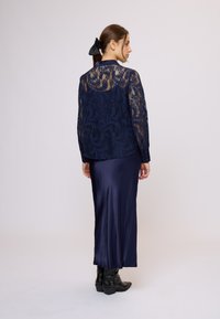 Tengerészkék csipkeblúz hosszú ujjakkal és virágmintákkal, selyem tengerészkék maxiszoknyával párosítva, fekete bokacsizmával kiegészítve.