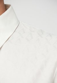 Hvit skjorte med subtilt preget geometrisk mønster og en spiss krage.