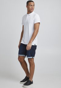 T-shirt polo blanche à manches courtes et col, associée à un short en jean foncé avec des ourlets retroussés et des baskets noires simples.