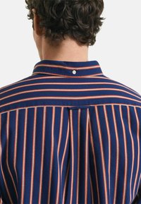 Chemise col montant avec des rayures verticales bleu marine et orange. Fabriquée dans un tissu à texture lisse, mise en valeur par un détail de bouton au dos.