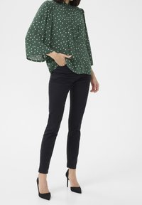 Blusa verde con pois bianchi, davanti arricciato, maniche lunghe e colletto alla mandarina, abbinata a pantaloni neri slim-fit e tacchi neri.