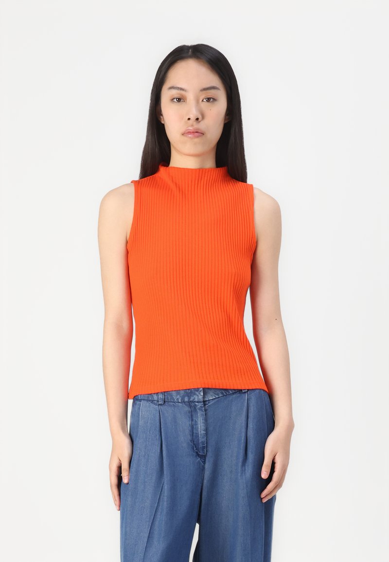 Top sans manches orange à côtes avec un col haut ; coupe ajustée, tissu léger, texture lisse, associé à un pantalon en denim ample.
