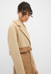 Beige uldblandingjakke med en struktureret krave, læderbælte detaljer og brede ærmer. Stylet med en matchende beige nederdel.