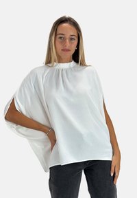 Top corto bianco a maniche corte con colletto alto, caratterizzato da dettagli arricciati e una vestibilità oversize, abbinato a jeans grigio scuro. Texture liscia.