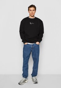Sort sweatshirt med hvid broderi, parret med blå denimjeans. Fodtøjet har et gråt og beige design. Enkel, afslappet outfit.