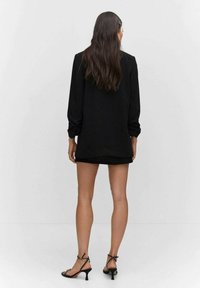 Nainen, jolla on pitkä ruskea hius ja päällä musta bleiserimekko sekä mustat hihnasandaalit korkoilla, seisoo selkä suorana tasaisen valkoisen seinän edessä.