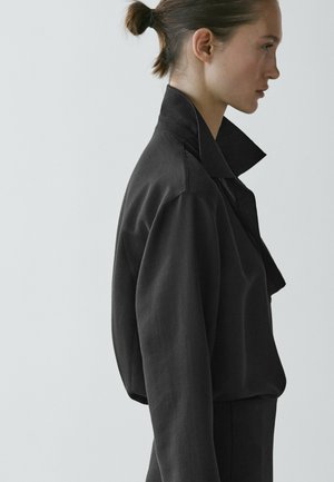 Donna con capelli scuri raccolti indossa una giacca nera oversize con collo alto, in piedi di profilo su uno sfondo chiaro e uniforme.