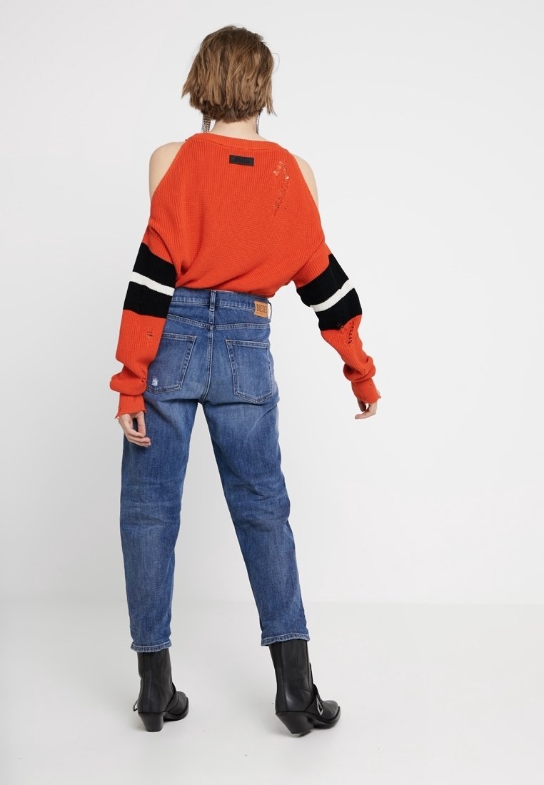 Pull en tricot rouge avec un design épaule nue, manches rayées noir et blanc ; associé à un jean taille haute bleu et des bottines noires.