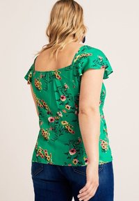 Grøn blomstret bluse med korte volangærmer, firkantet udskæring og løst fit, parret med mørkeblå jeans.
