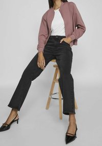 Woman seated on wooden stool wearing mauve zip jacket, white top, black high-waist jeans, and black pointed slingback heels.