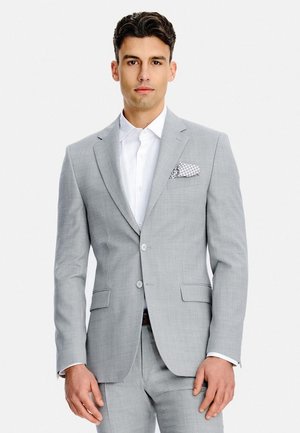 Mężczyzna w jasnoszarym garniturze z białą koszulą i wzorzystą poszetką, stojący na jednolitym tle.