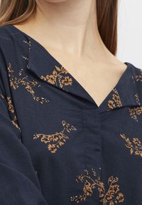 Nærbillede af en person iført en mørkeblå top med brune blomsterprint og en bred, foldet krave.