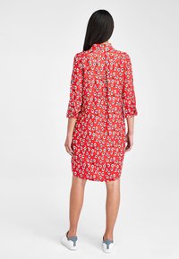 Robe rouge à taches irrégulières noires et blanches, manches trois-quarts, dos boutonné, associée à des baskets blanches, texture de tissu lisse.