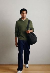 Felpa verde con cappuccio, jeans blu scuro e sneakers bianche. Borsa a tracolla nera. Modella che cammina su un pavimento di legno davanti a una parete grigio chiaro.