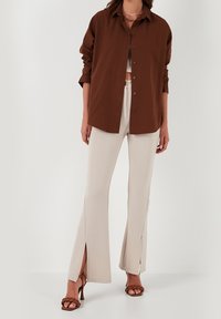 Donna che indossa una camicia marrone con bottoni sopra pantaloni beige a palazzo con fessure anteriori e sandali alti intrecciati marroni.