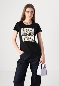 Camiseta negra con un parche cuadrado que presenta patrones abstractos en dorado, blanco y negro, y "LIUJO" impreso en letras negritas negras.