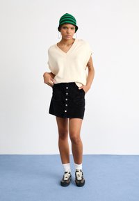 Pull beige en maille à col en V, mini-jupe en velours côtelé noir avec boutons sur le devant, bob rayé vert et noir, baskets décontractées, chaussettes blanches.