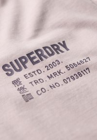Lyserød bomulds-T-shirt med mørkegrå tekst, der viser mærket "SUPERDRY", etableringsdato og produktdetaljer. Blød stoftekstur.