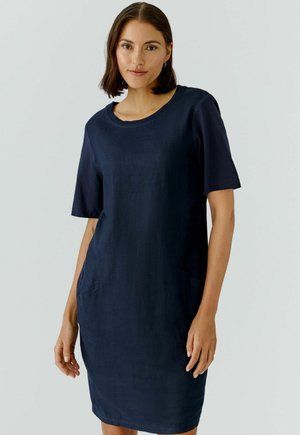 Femme debout portant une robe bleu marine à manches courtes, longueur genou, col rond et poches latérales, fond neutre.