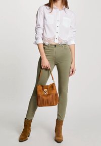 Bílá zkrácená košile s krajkovým lemem, olivově zelené slim fit kalhoty, hnědé semišové kotníkové boty a hnědá kabelka s třásněmi.