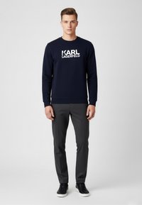 Navy Sweatshirt mit großem weißen "KARL LAGERFELD" Logo, kombiniert mit grauen Hosen und schwarzen Sneakers. Einfaches, modernes Design und Passform.