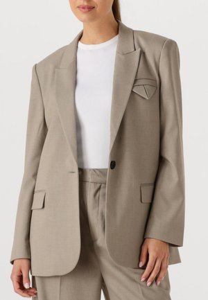 Woman wearing a beige blazer and matching trousers over a white ribbed crew neck top, standing against a plain background.